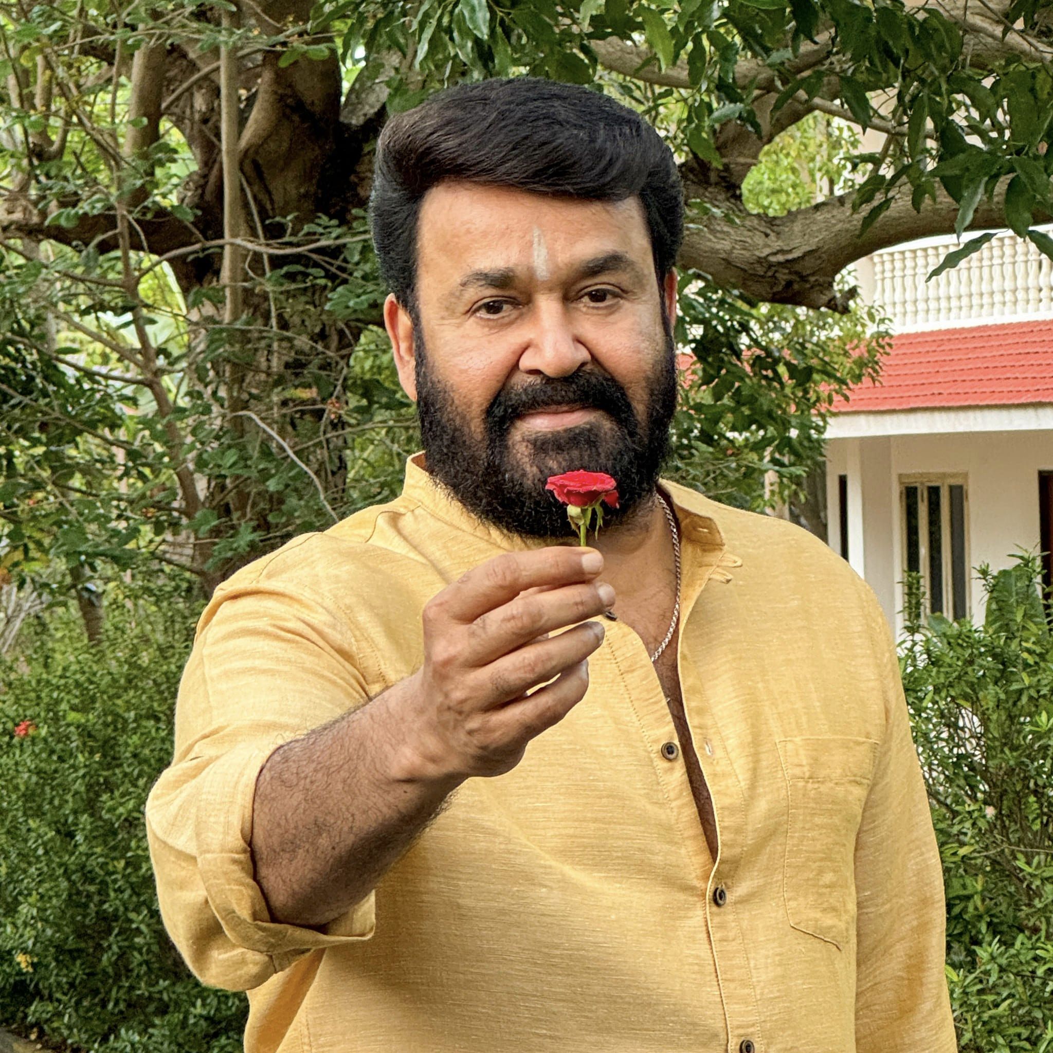 Mohanlal standing in a garden. He is shown from the waist up wearing a yellow shirt and extending a red flower to the user.